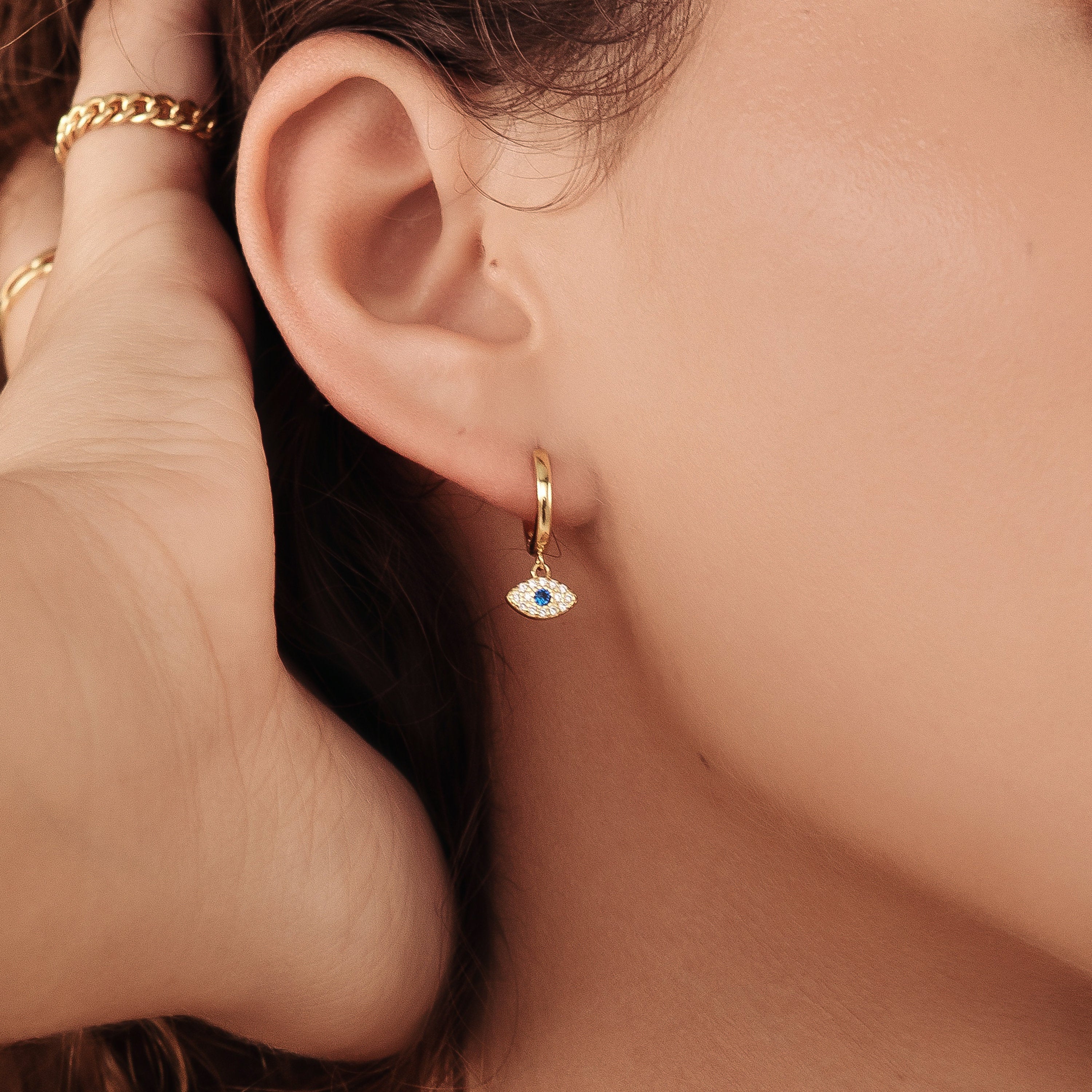 Close-up of a woman's ear wearing the Iris Huggies—gold huggie earrings with a small blue and white evil eye charm, perfect for adding stylish protection to your jewelry collection.