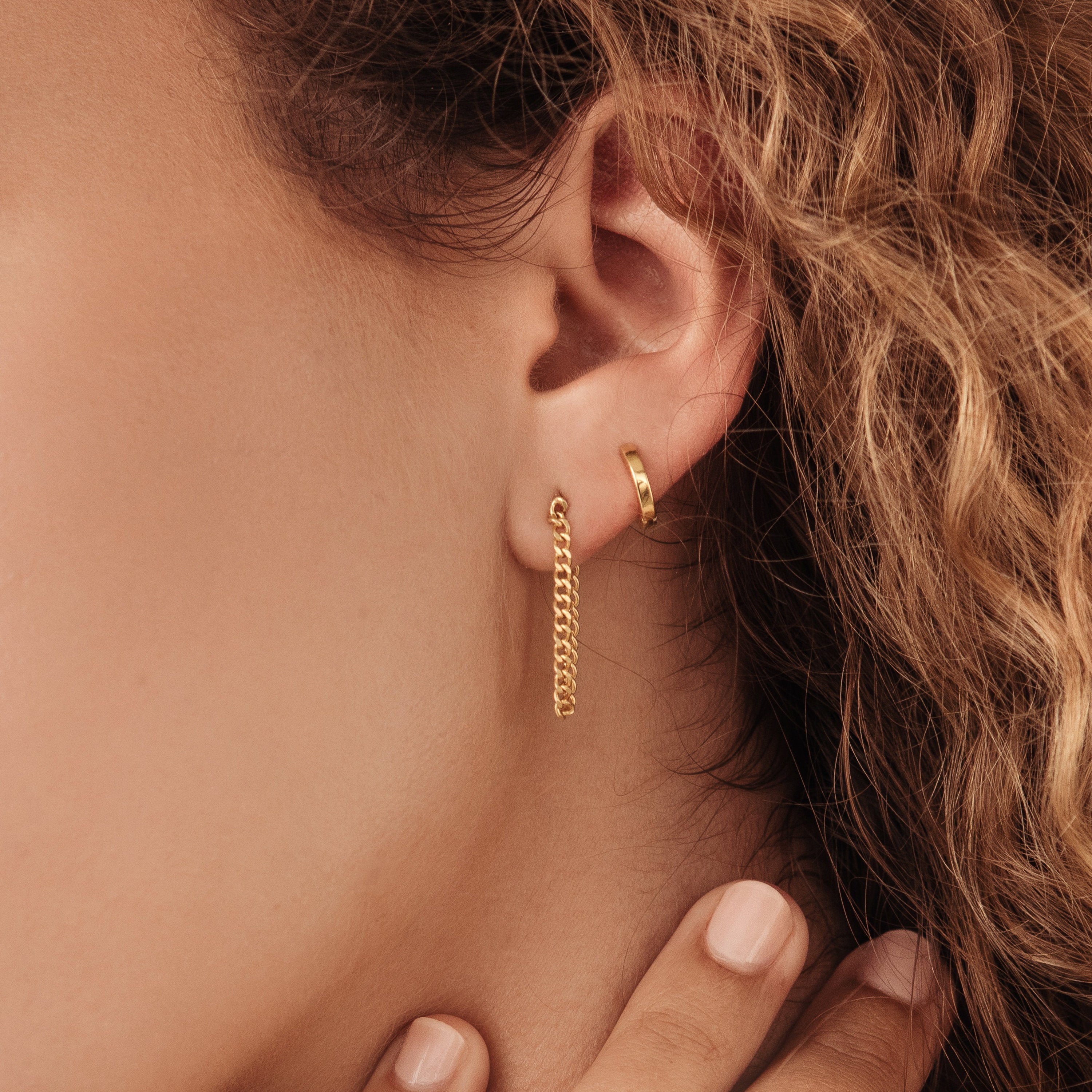 Close-up of a woman's ear wearing the Editor Huggies—one gold hoop and one chain earring—framed by curly hair for a minimal, classic look.