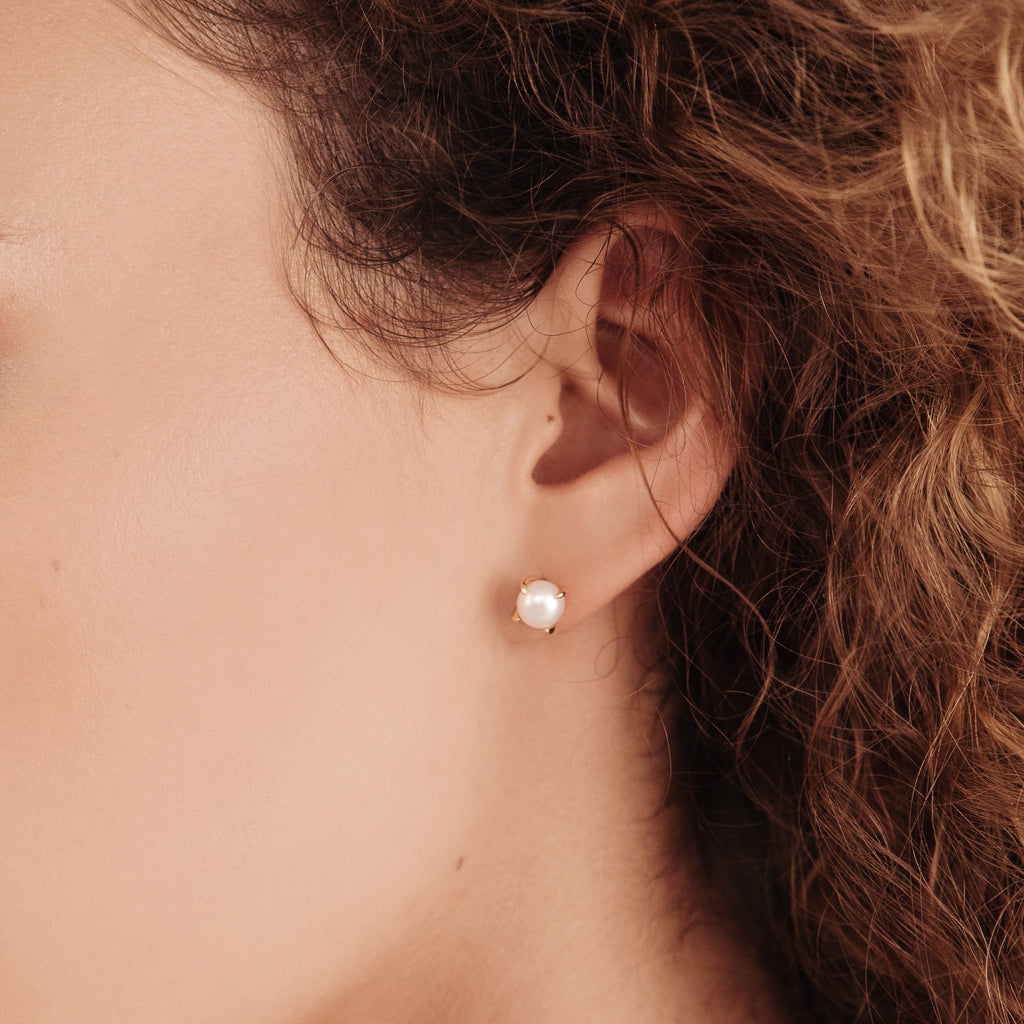 A close-up of a woman’s ear wearing Zoe Pearl Studs—small, round pearl earrings that add an elegant touch to her look—framed by her curly brown hair.