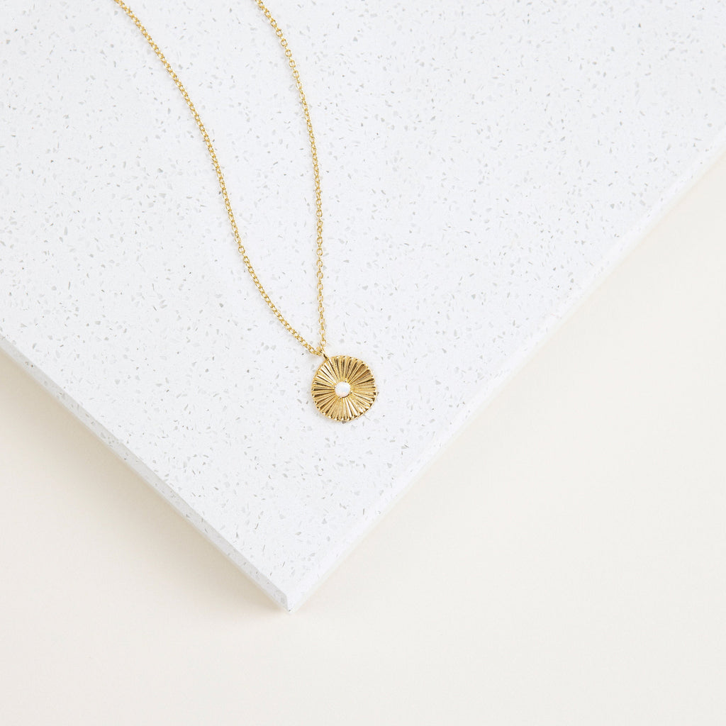 The Nova Necklace features a sunburst design with a center gem, inspired by vintage jewelry, and is displayed on a white speckled surface.