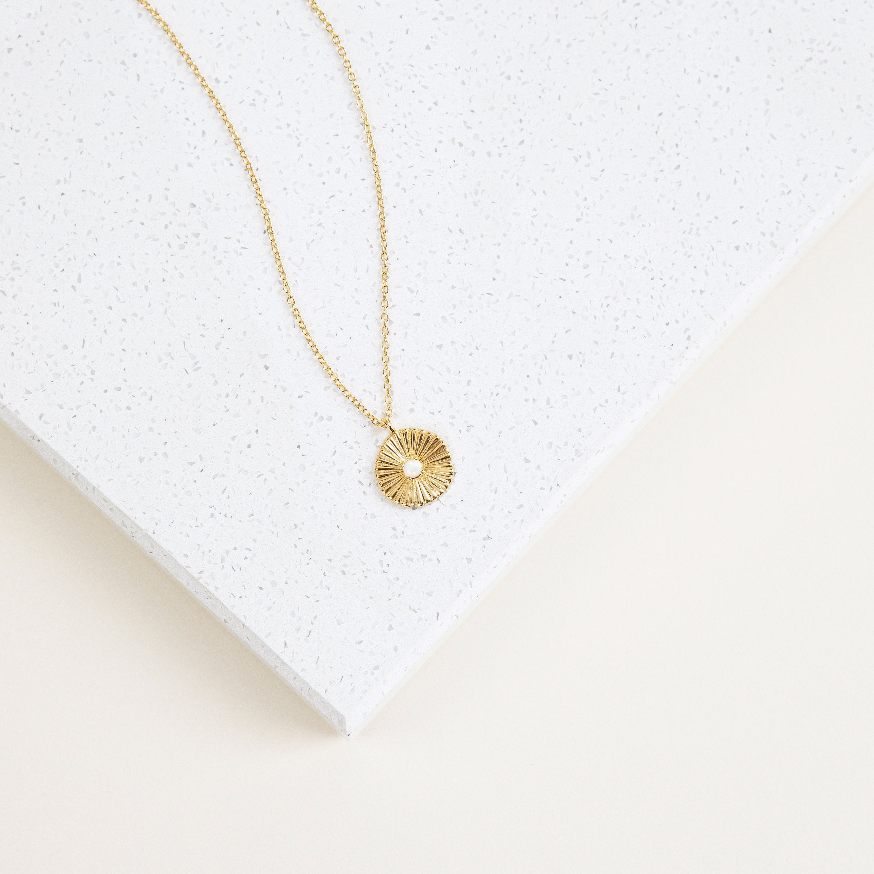 The Nova Necklace features a sunburst design with a center gem, inspired by vintage jewelry, and is displayed on a white speckled surface.