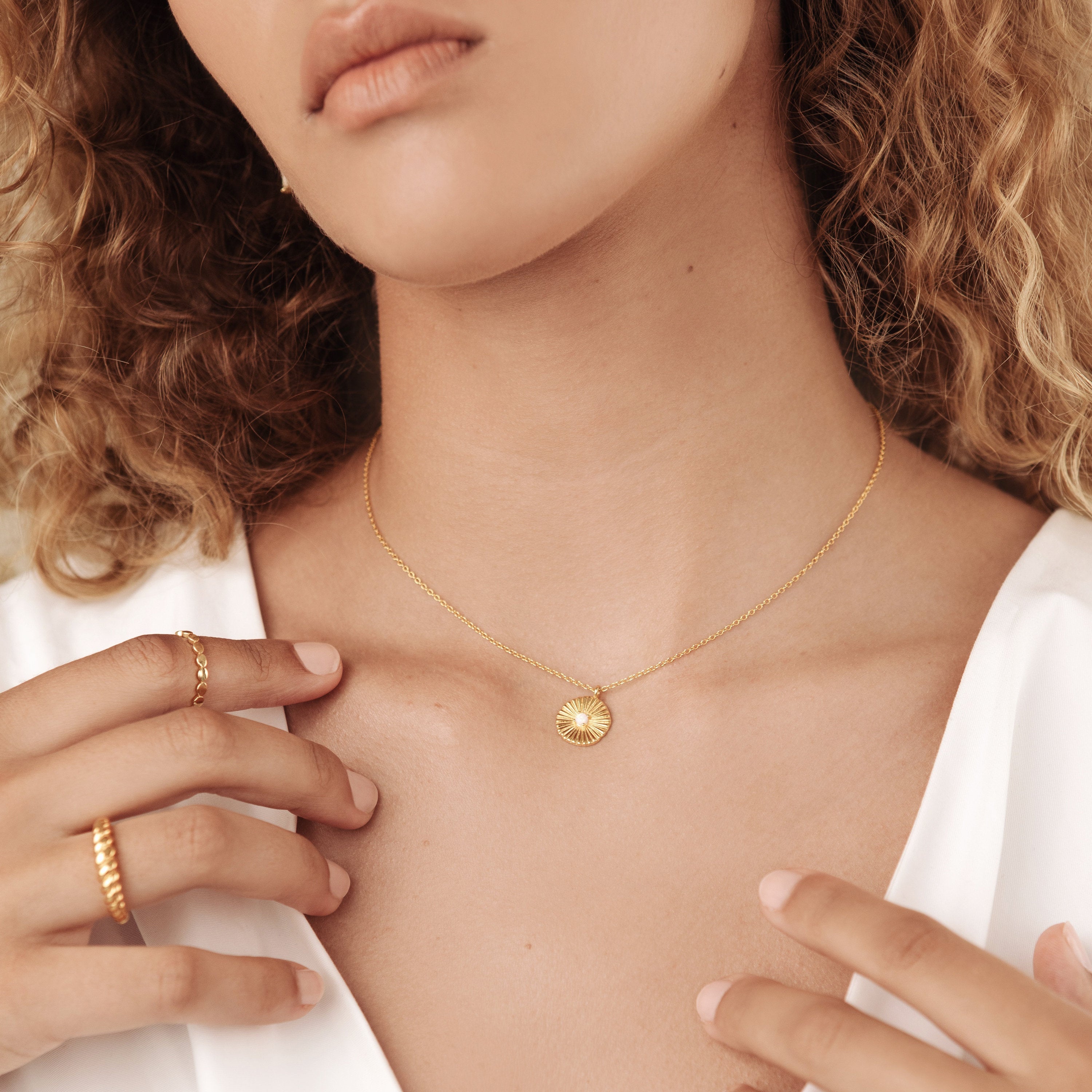 A woman in a white top wears the Nova Necklace with its round pendant and a gold ring—an elegant touch of vintage-inspired jewelry.