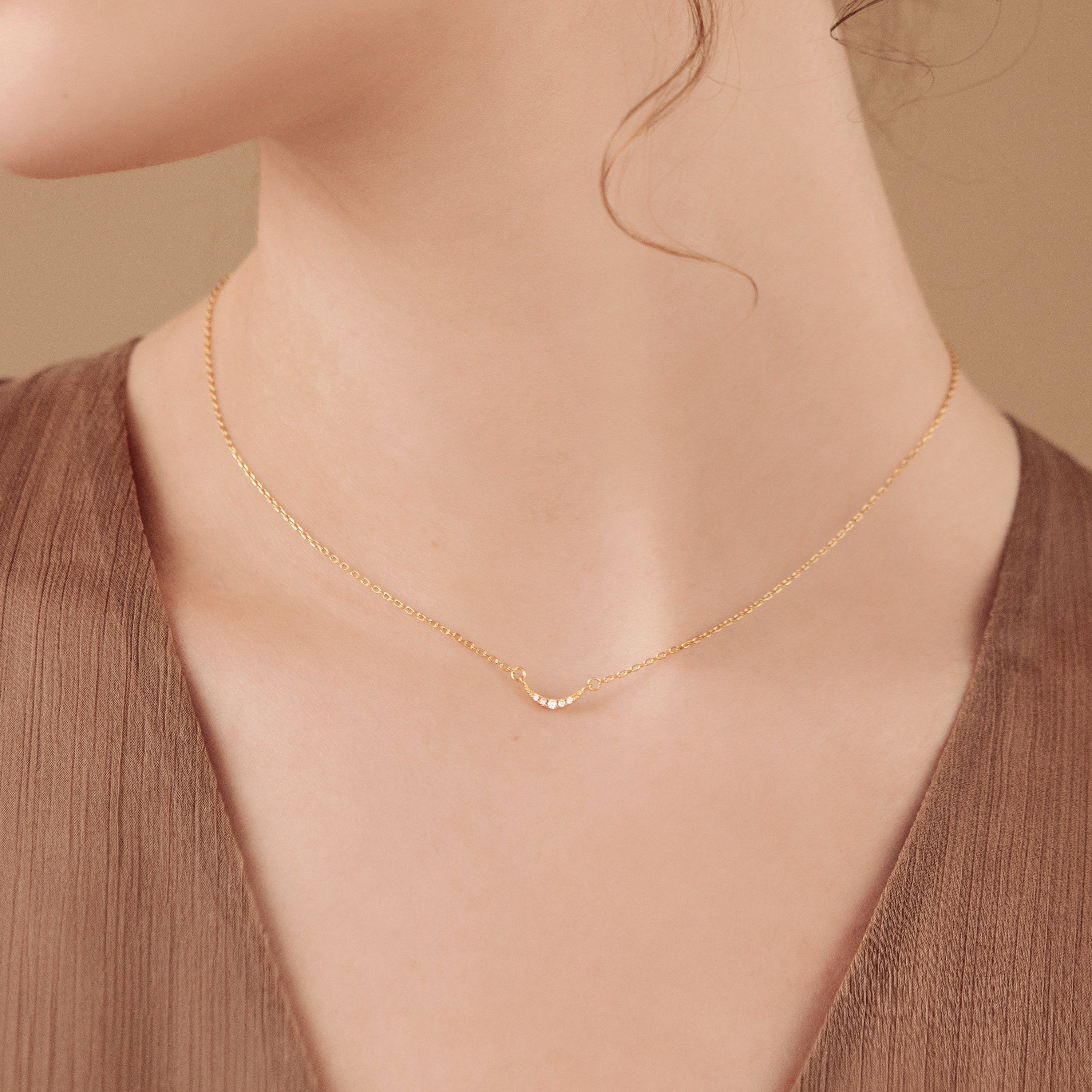 A woman wears the Lucia Pave Necklace, featuring a delicate crescent charm pendant, paired with a brown V-neck top.
