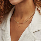 Close up of a woman wearing a layered gold necklace featuring a beaded chain with polished spheres and a delicate chain with a single gold bead pendant, worn with a textured white collared top for a modern, elegant look.