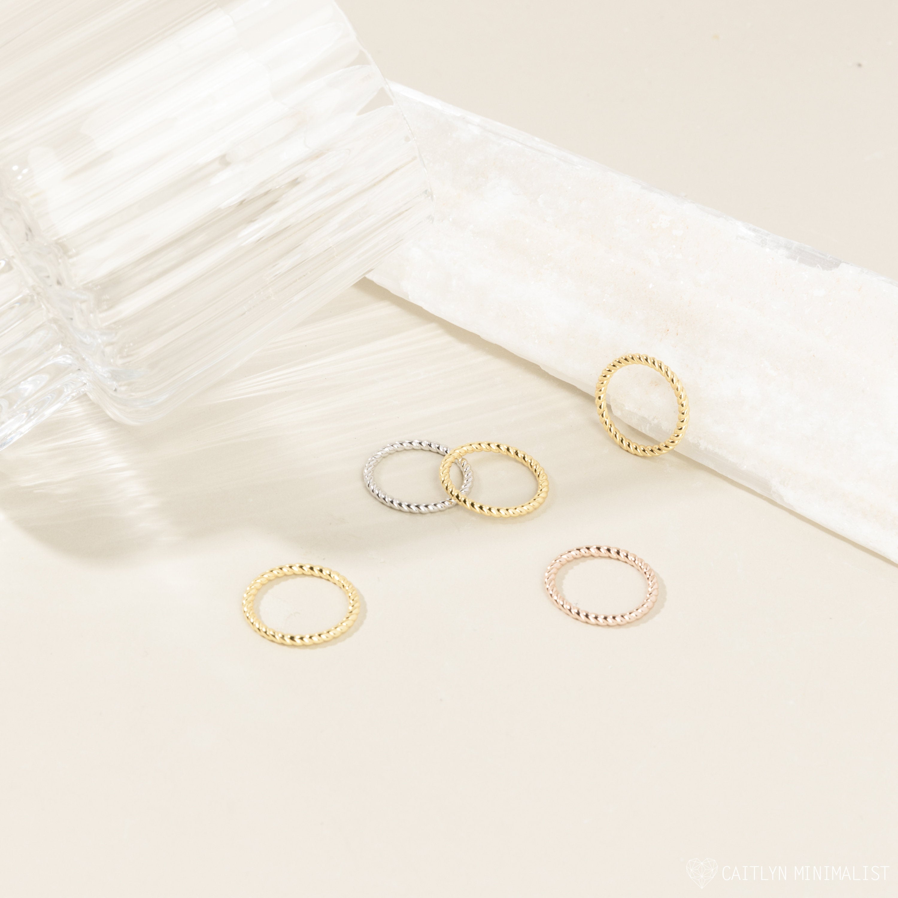Five textured Lea Twisted Rings in gold, silver, and rose gold, including twisted and layered designs, are displayed on a light surface beside a glass and a white object.