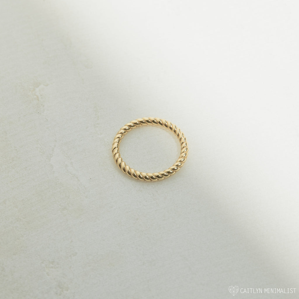 The Lea Twisted Ring, featuring polished gold bands in a simple twisted design, rests on a lightly textured surface with soft shadows.