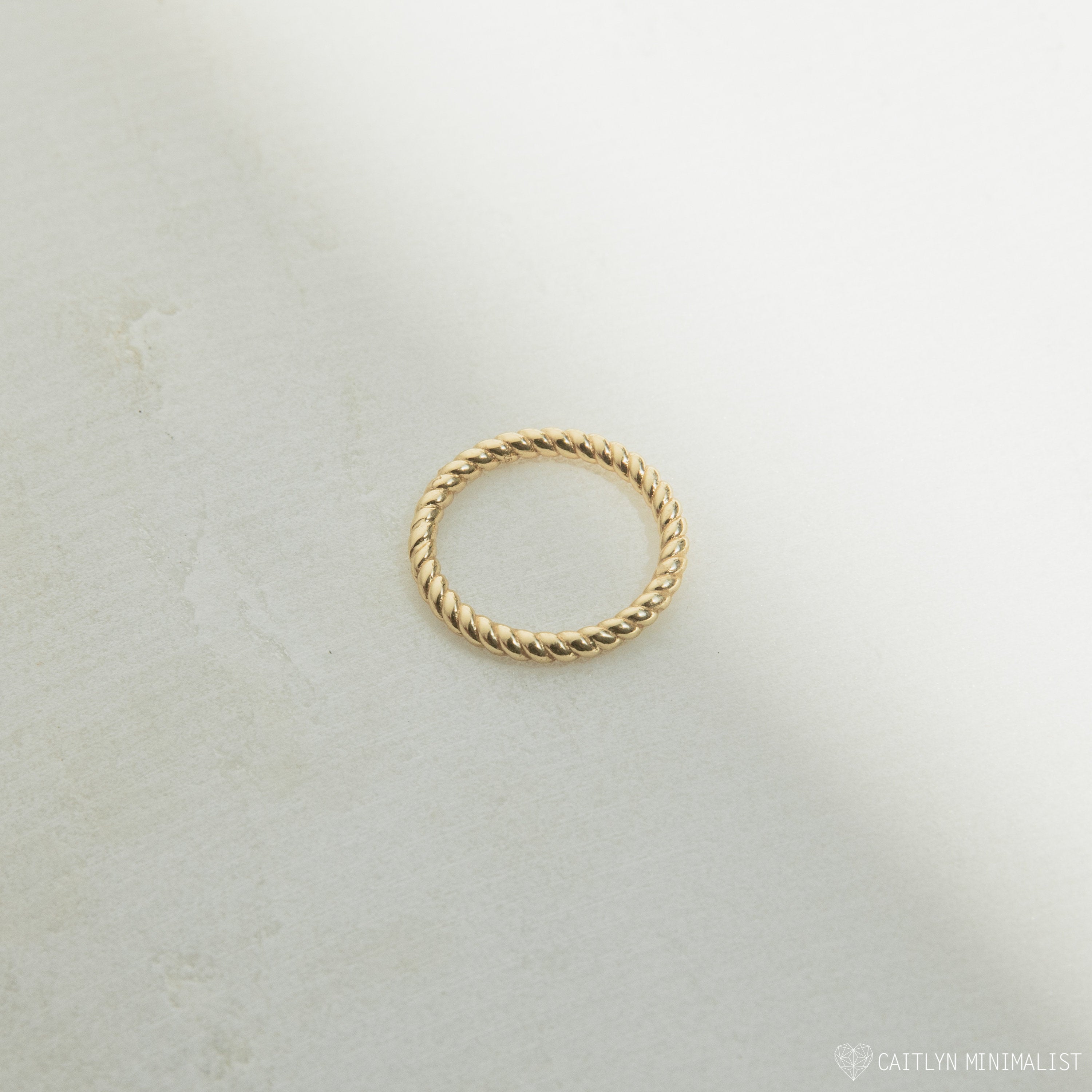 The Lea Twisted Ring, featuring polished gold bands in a simple twisted design, rests on a lightly textured surface with soft shadows.