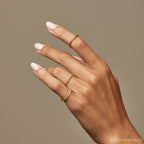 A hand with light pink nails wears three thin gold rings, including the Lea Twisted Ring, against a beige background.