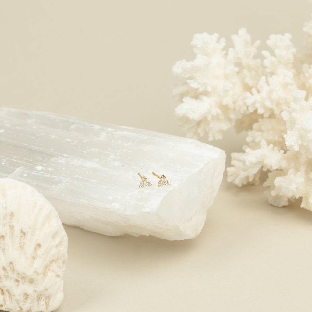 Mini Lily Studs in Sterling Silver are displayed on a white crystal, with coral and a shell in the background—perfect for everyday wear or adding sparkle to cartilage piercings.