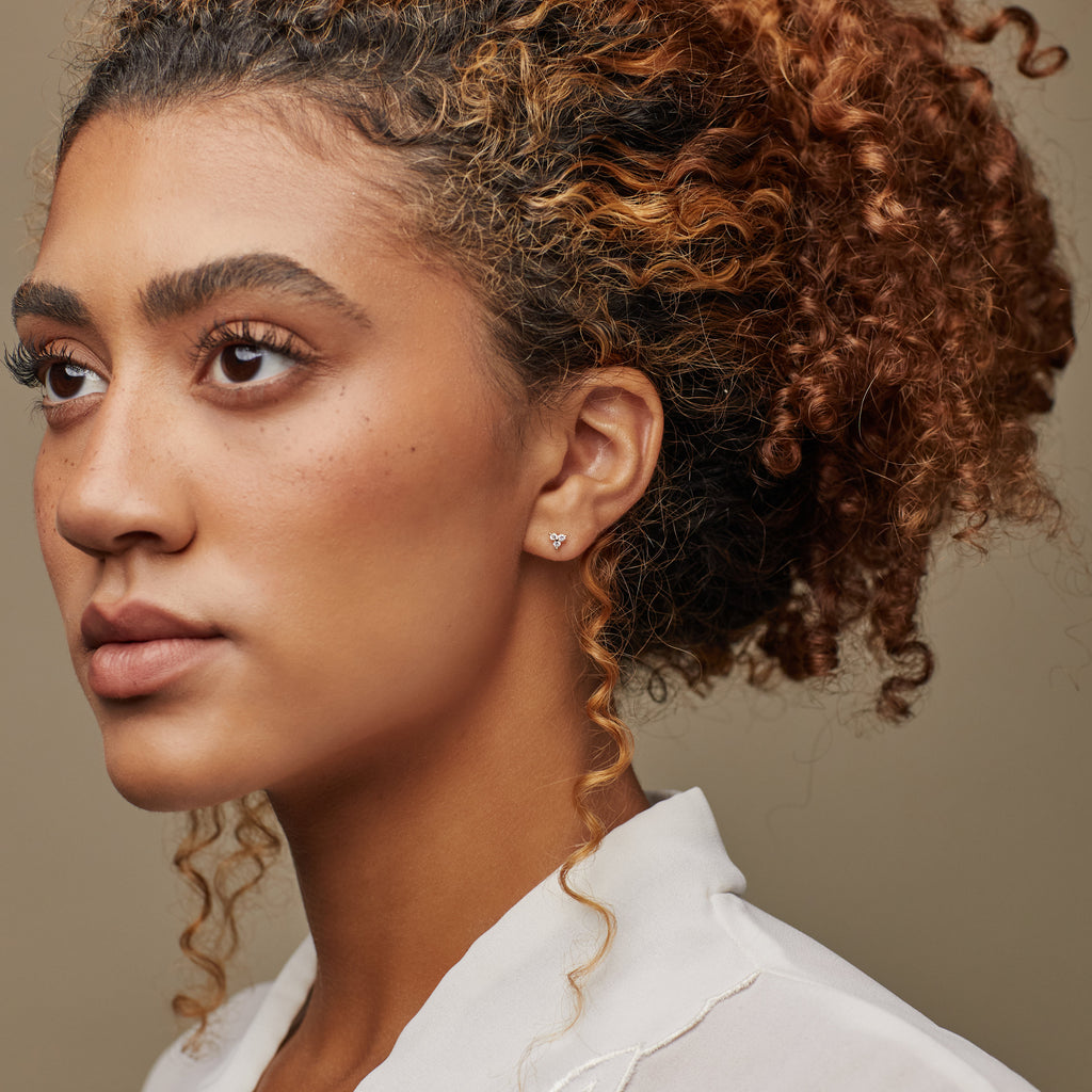 A woman with curly hair wearing a white blouse and Mini Lily Studs, the perfect everyday earrings, plus a small heart-shaped cartilage earring, looks to the left.