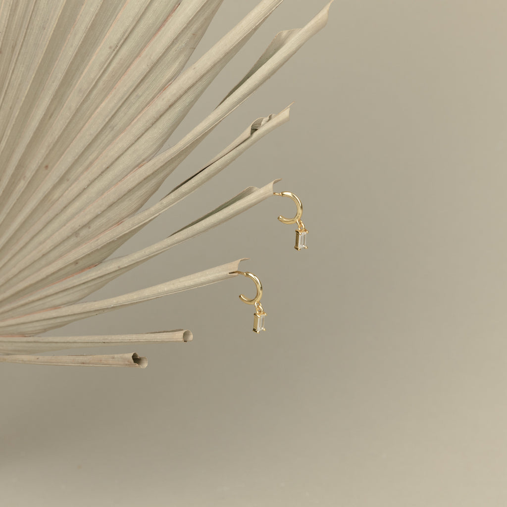 Lys Baguette Hoops, featuring small charms and baguette diamonds, exude classic elegance and are displayed on a dried palm leaf with a beige background.