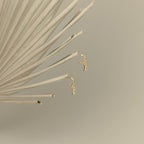 Lys Baguette Hoops, featuring small charms and baguette diamonds, exude classic elegance and are displayed on a dried palm leaf with a beige background.