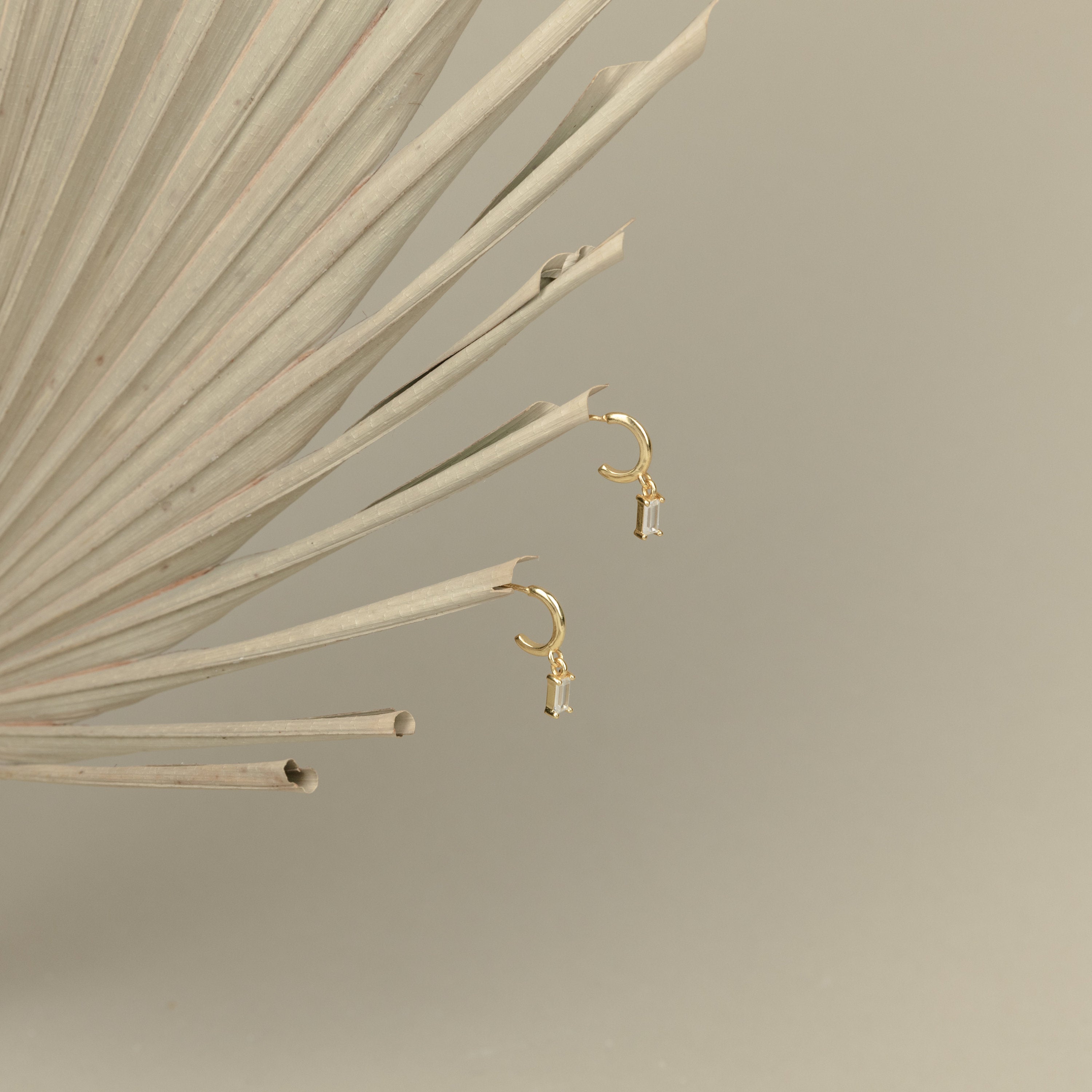 Lys Baguette Hoops, featuring small charms and baguette diamonds, exude classic elegance and are displayed on a dried palm leaf with a beige background.
