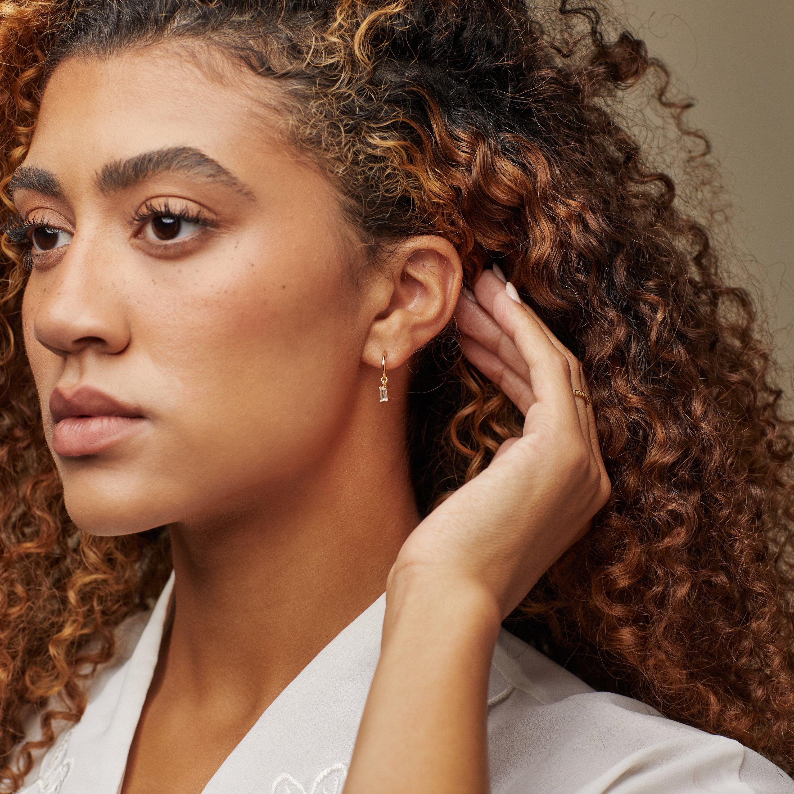 A woman with curly hair, hand on ear, exudes classic elegance while wearing Lys Baguette Hoops.