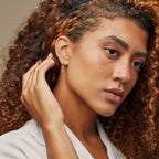 A woman with curly hair in a white top touches her ear, wearing Lys Baguette Hoops and radiating classic elegance.
