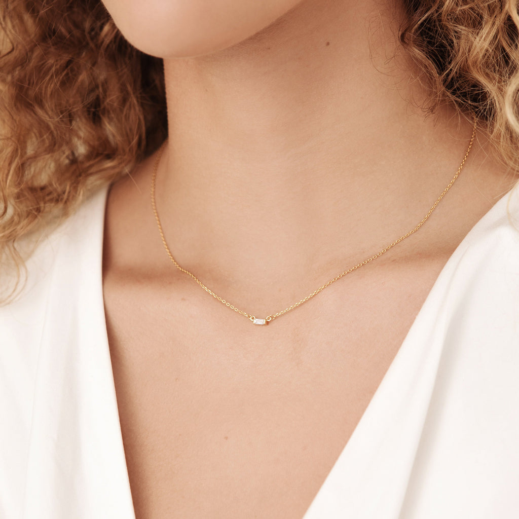 A woman wears the Emery Necklace in Sterling Silver—a minimal piece with a delicate chain and tiny pendant—paired with a white top. This necklace makes a perfect holiday gift.
