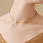 Close-up of a woman wearing two gold chain necklaces, including the Skyy Toggle Necklace, against a beige background. Ideal for minimal jewelry lovers.