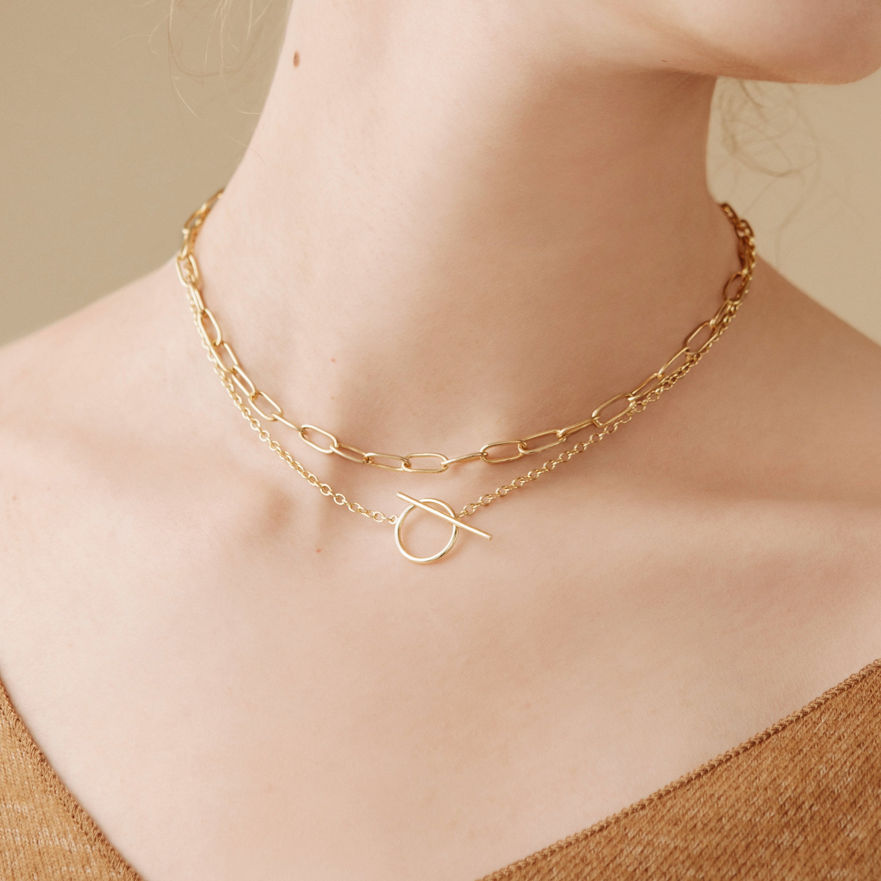 Close-up of a woman wearing two gold chain necklaces, including the Skyy Toggle Necklace, against a beige background. Ideal for minimal jewelry lovers.