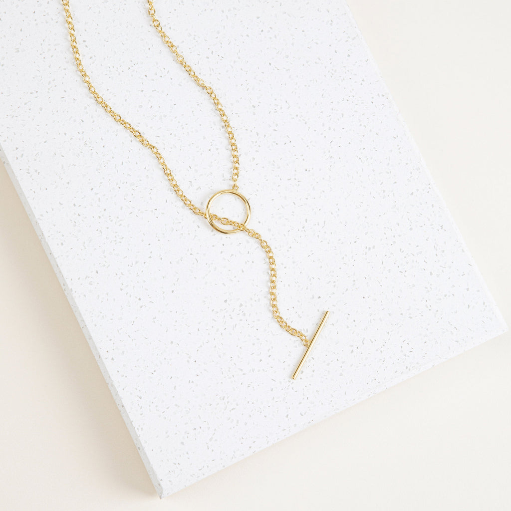 The Skyy Toggle Necklace features a gold bar pendant, ideal for minimal jewelry lovers. Shown on a textured white rectangular surface.