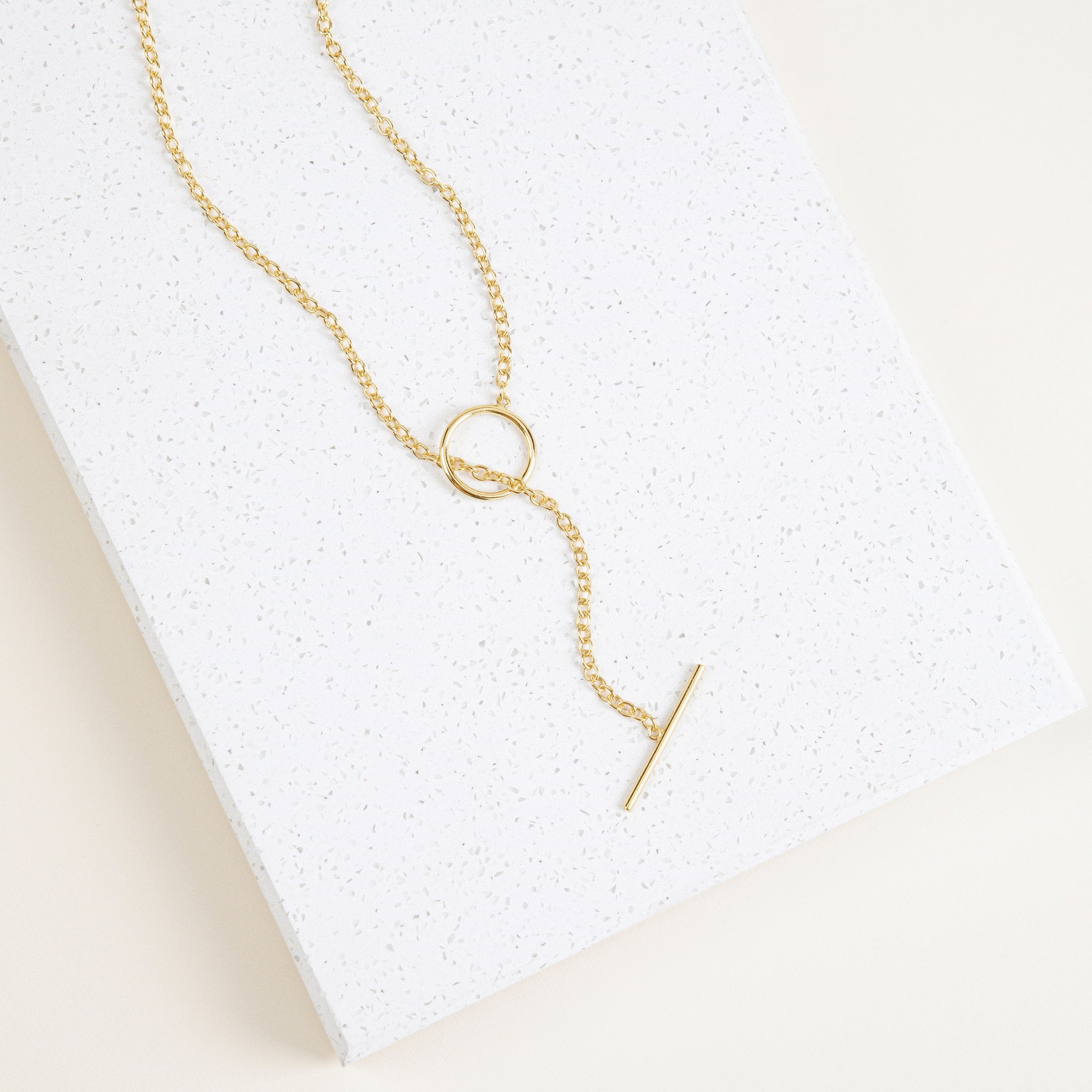 The Skyy Toggle Necklace features a gold bar pendant, ideal for minimal jewelry lovers. Shown on a textured white rectangular surface.