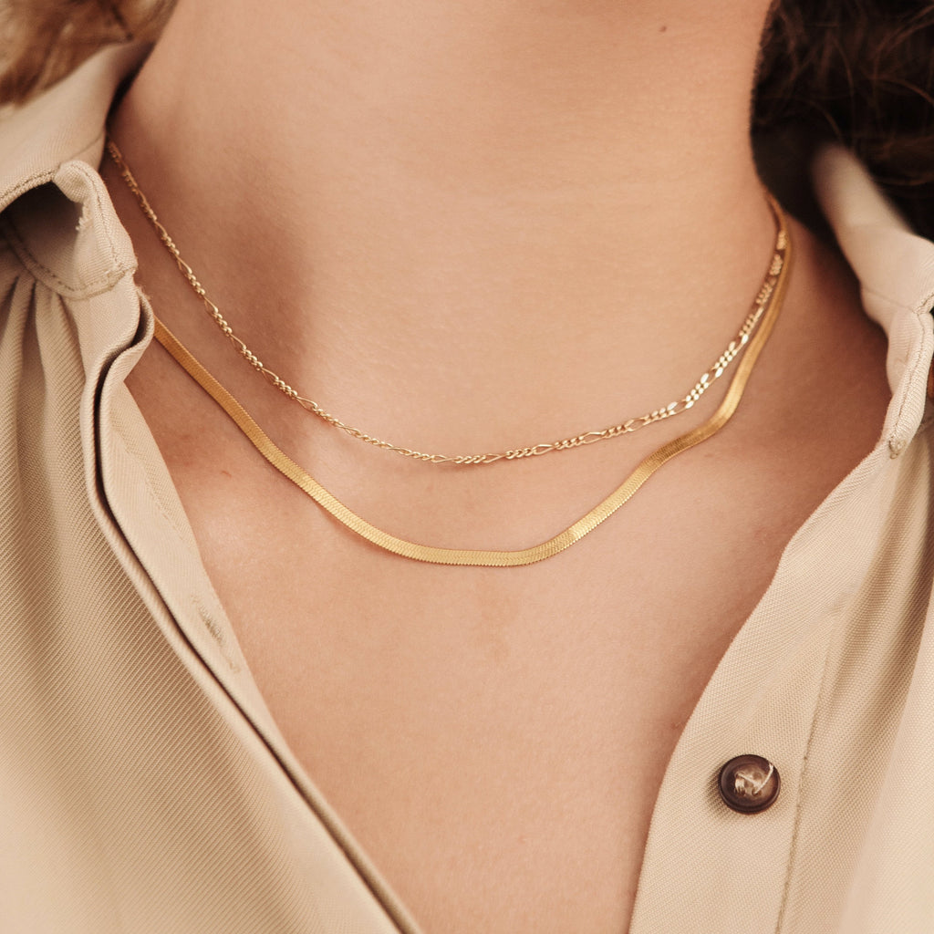 Two necklaces layered on a model: the Figaro necklace and a flat snake chain, styled with a neutral button-down shirt.