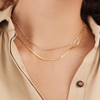 Two necklaces layered on a model: the Figaro necklace and a flat snake chain, styled with a neutral button-down shirt.