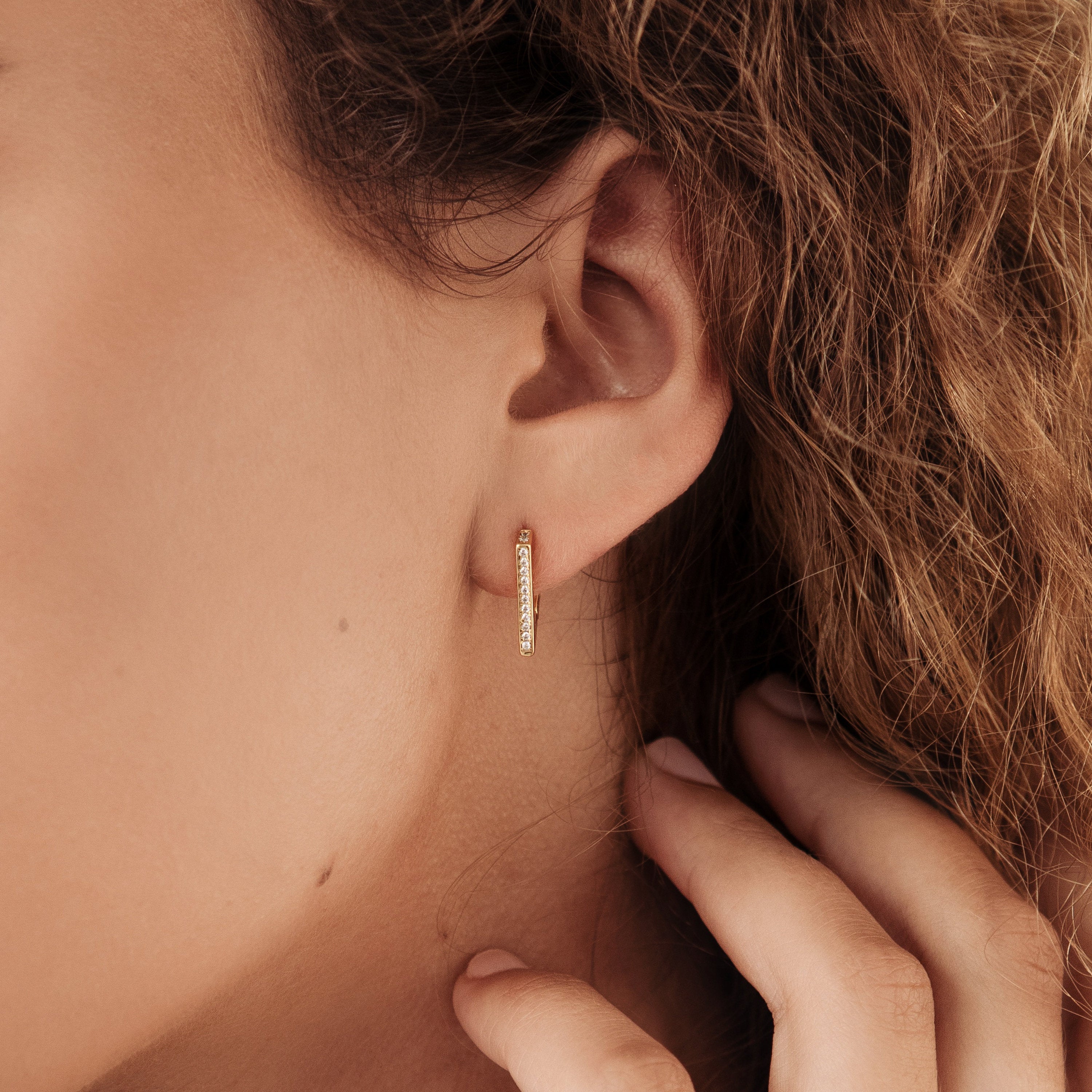 A close-up of a woman's ear wearing the Ezra Triangle Earrings—small gold hoops with pave texture—as her hand gently touches her neck, showcasing modern jewelry elegance.