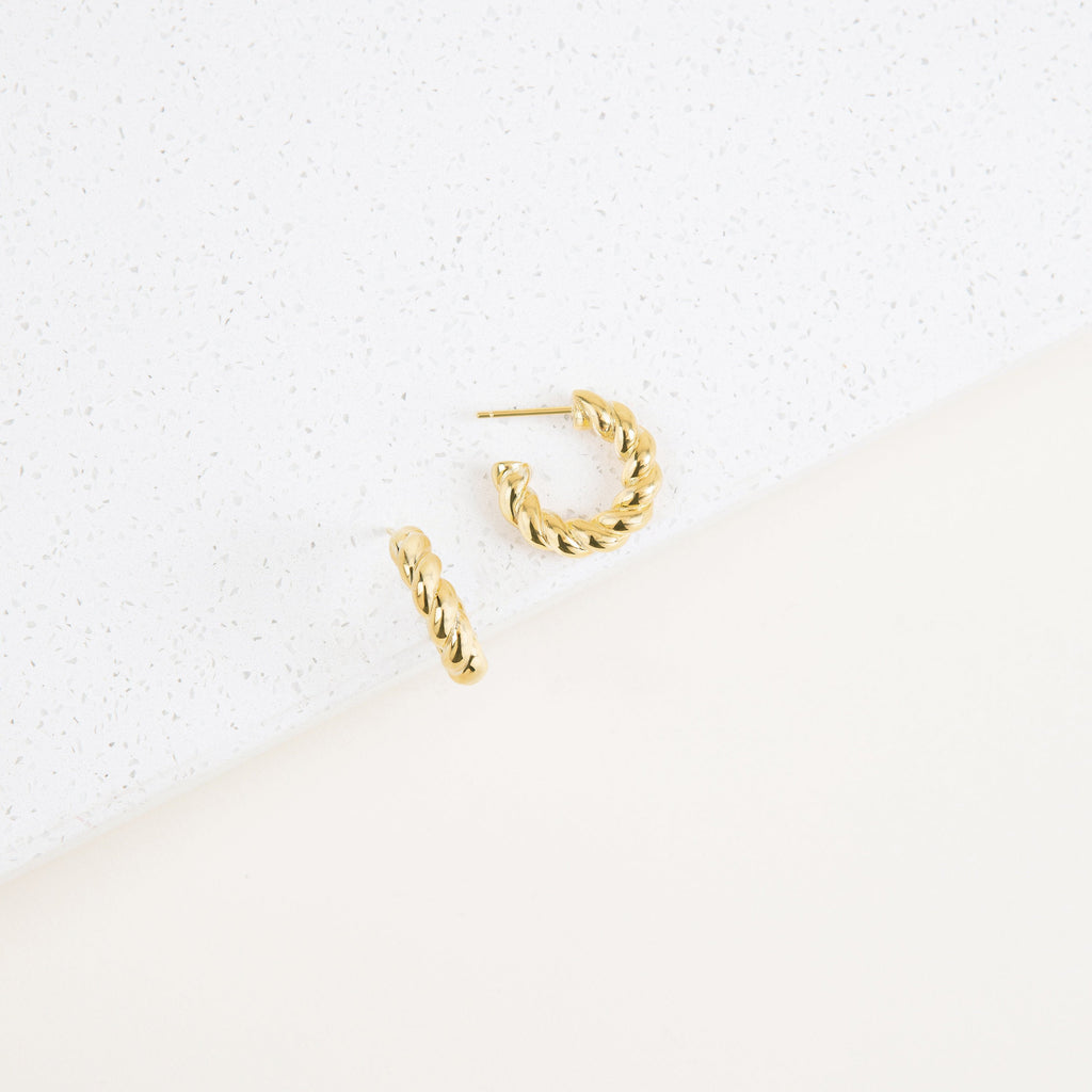 The Remi Twist Hoops, a pair of gold earrings, are displayed on a white textured surface with a minimalist background—perfect for fans of hoop earrings and minimal jewelry.