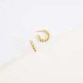 The Remi Twist Hoops, a pair of gold earrings, are displayed on a white textured surface with a minimalist background—perfect for fans of hoop earrings and minimal jewelry.
