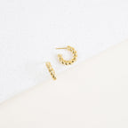 The Remi Twist Hoops, a pair of gold earrings, are displayed on a white textured surface with a minimalist background—perfect for fans of hoop earrings and minimal jewelry.