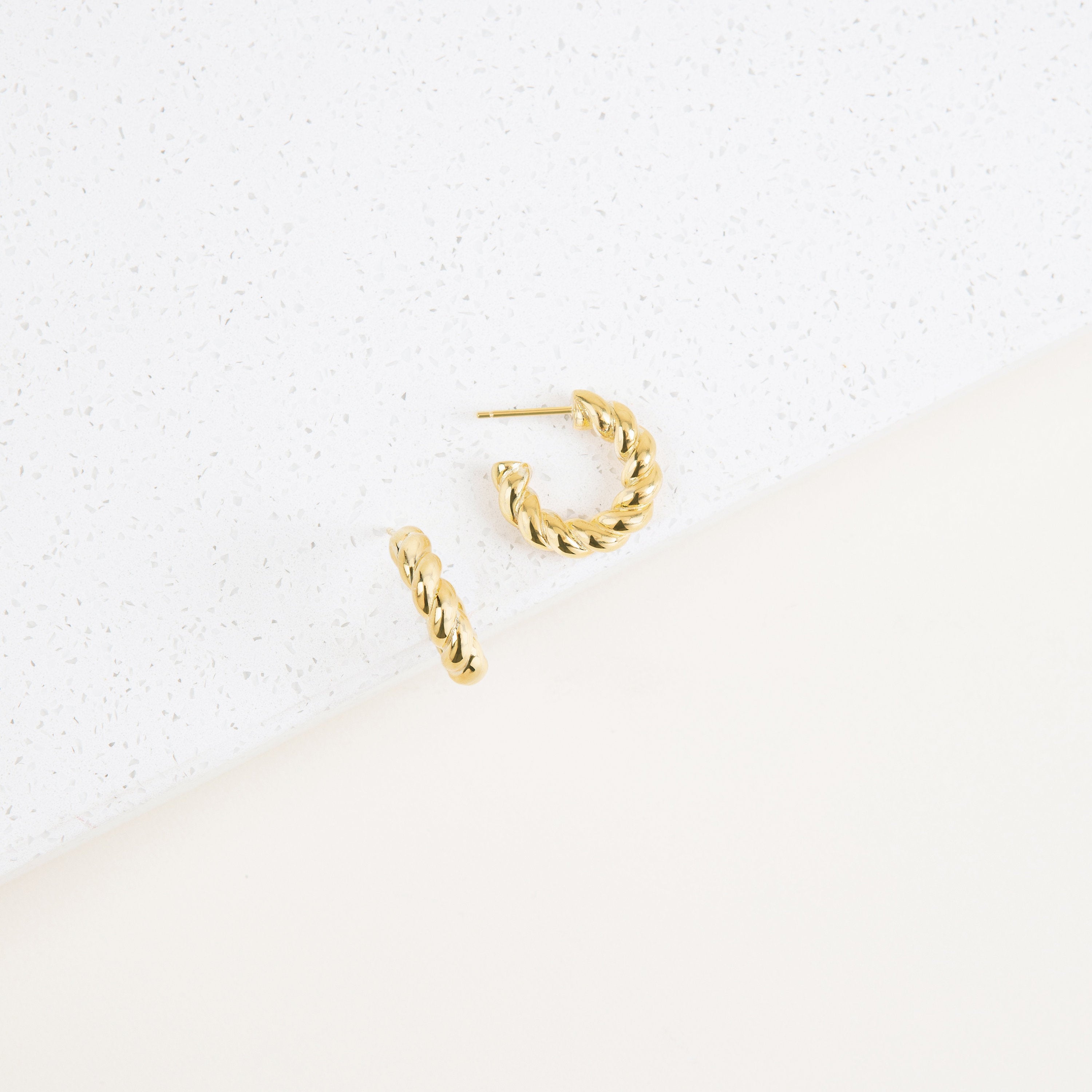 The Remi Twist Hoops, a pair of gold earrings, are displayed on a white textured surface with a minimalist background—perfect for fans of hoop earrings and minimal jewelry.
