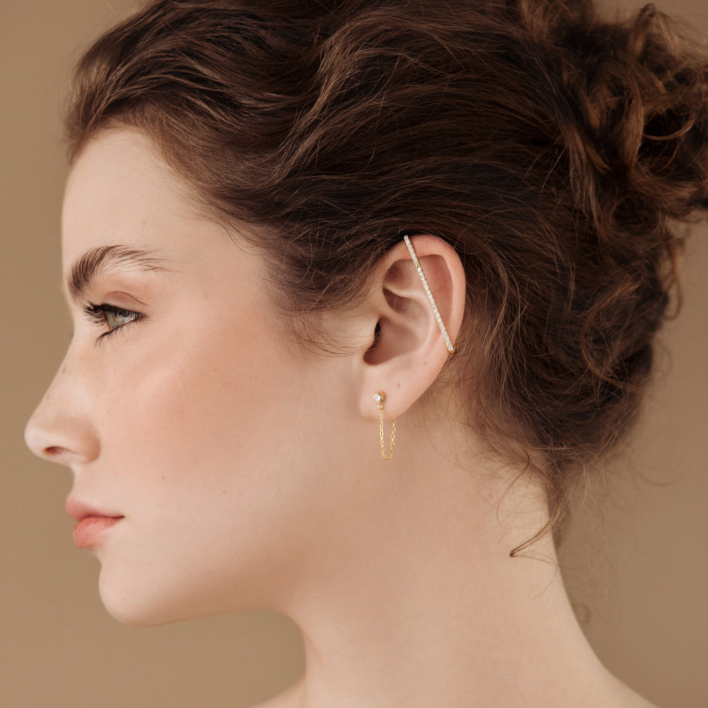 Profile of a woman with curly brown hair, wearing gold Bar Ear Cuffs and pave bar earrings, on a beige background.