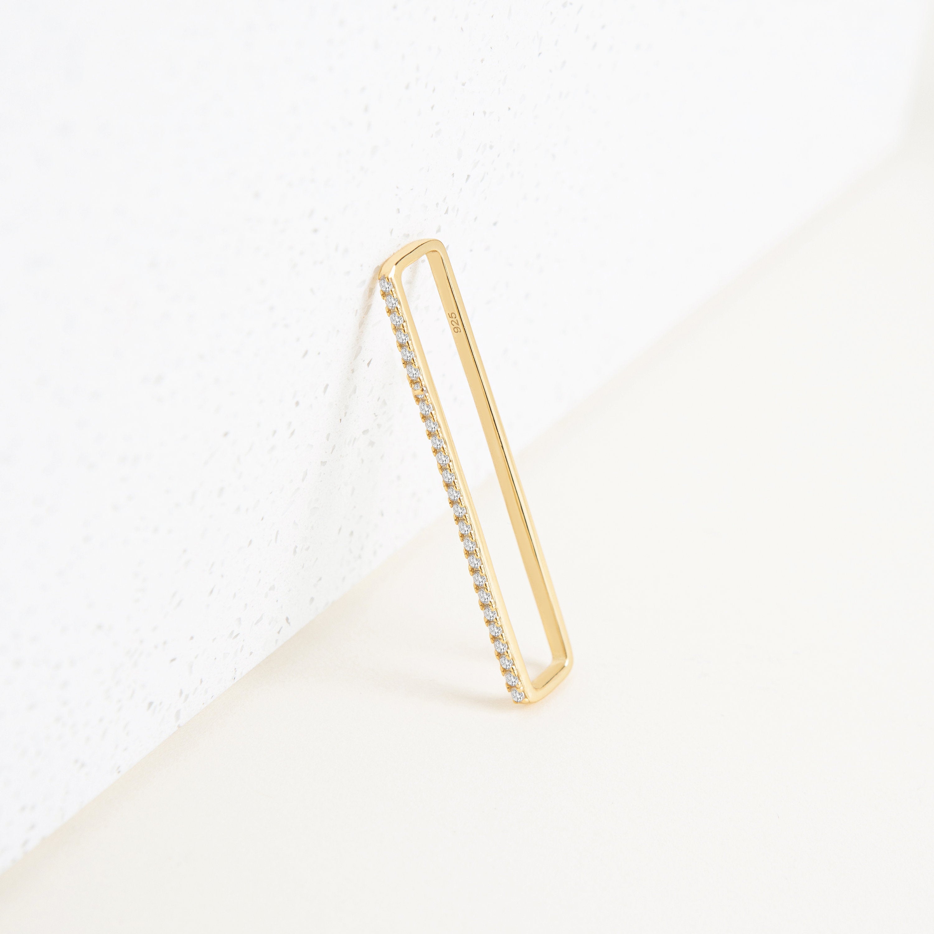 A pair of Bar Ear Cuffs, rectangular in shape with small embedded diamonds, reminiscent of pave jewelry, are leaning against a white textured surface.