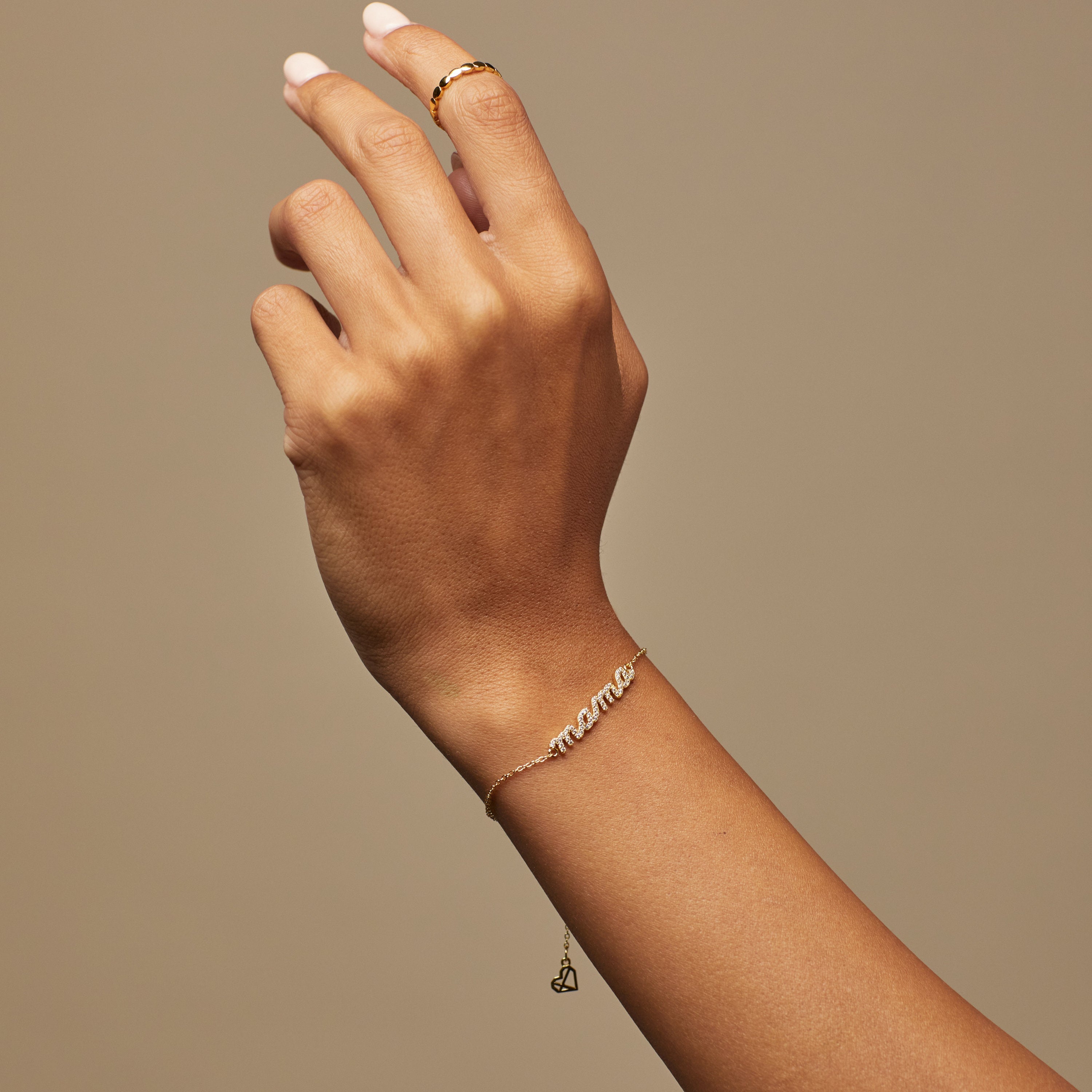 A hand adorned with a gold ring and the Pave Mama Script Bracelet, featuring the word "mama," gleams against a neutral background—an elegant choice for celebrating mama jewelry.