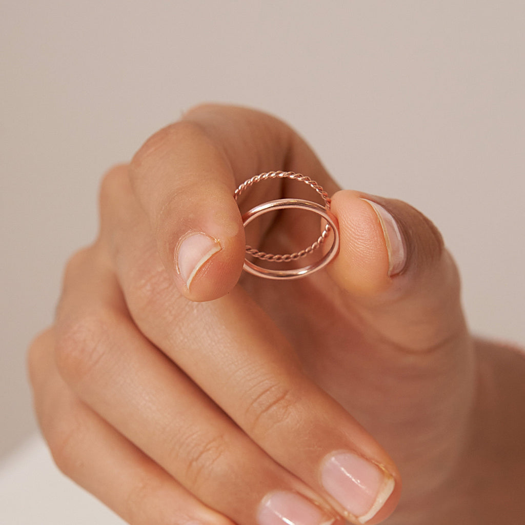 A hand displays the Minimalist Stacking Ring Set—two delicate, rose gold intertwined rings—against a neutral background, highlighting their minimalist design.