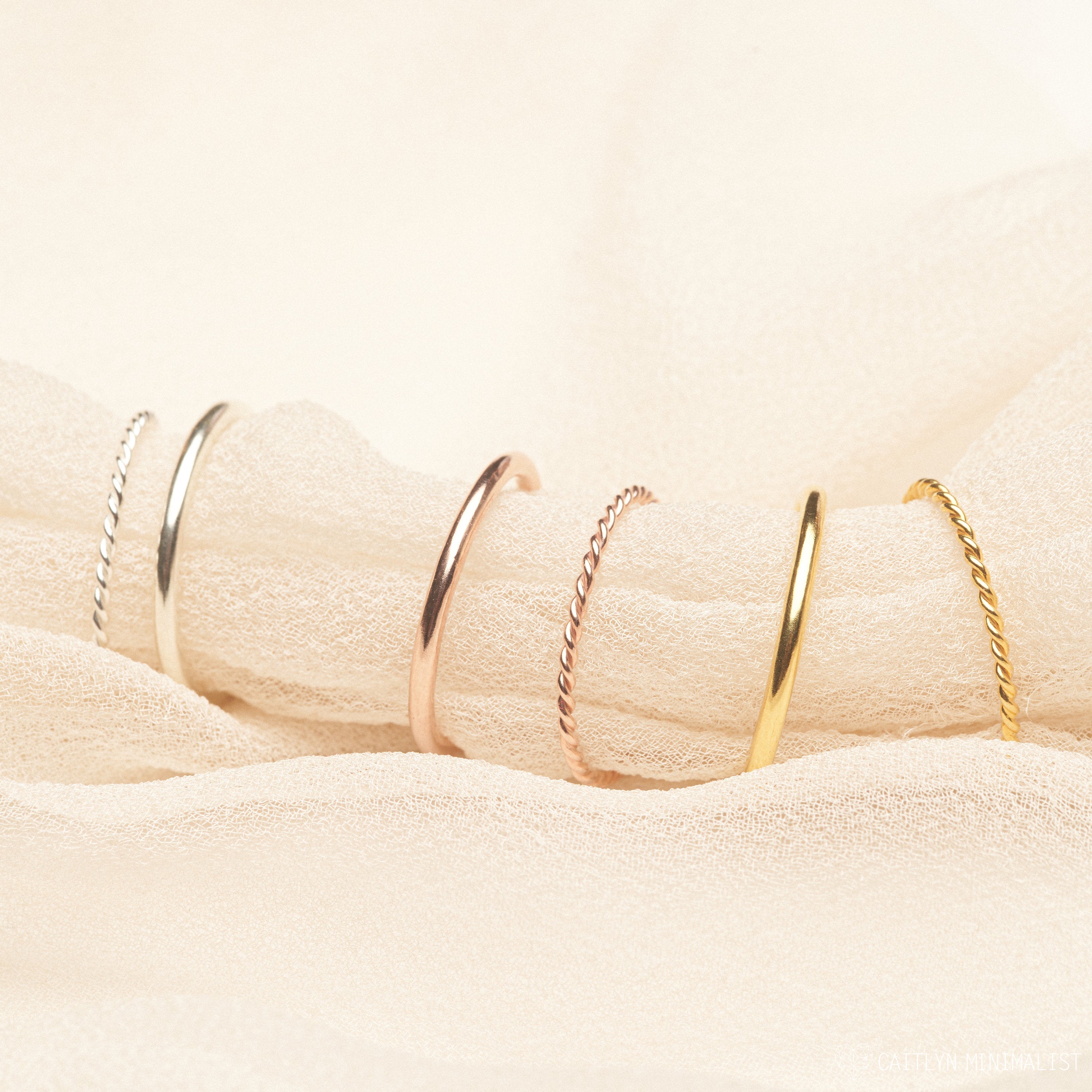 The Minimalist Stacking Ring Set features five delicate rings in silver, rose gold, and gold, elegantly displayed on soft cream fabric—perfect for refined minimalist style.