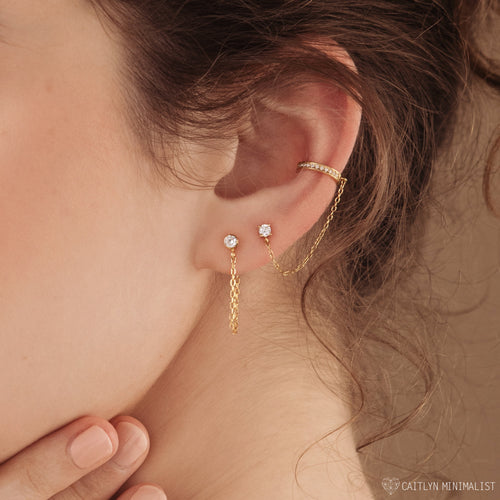 A close-up of an ear adorned with multiple gold earrings, including the versatile Birthstone Chain Studs featuring a chain that connects studs and a cuff.