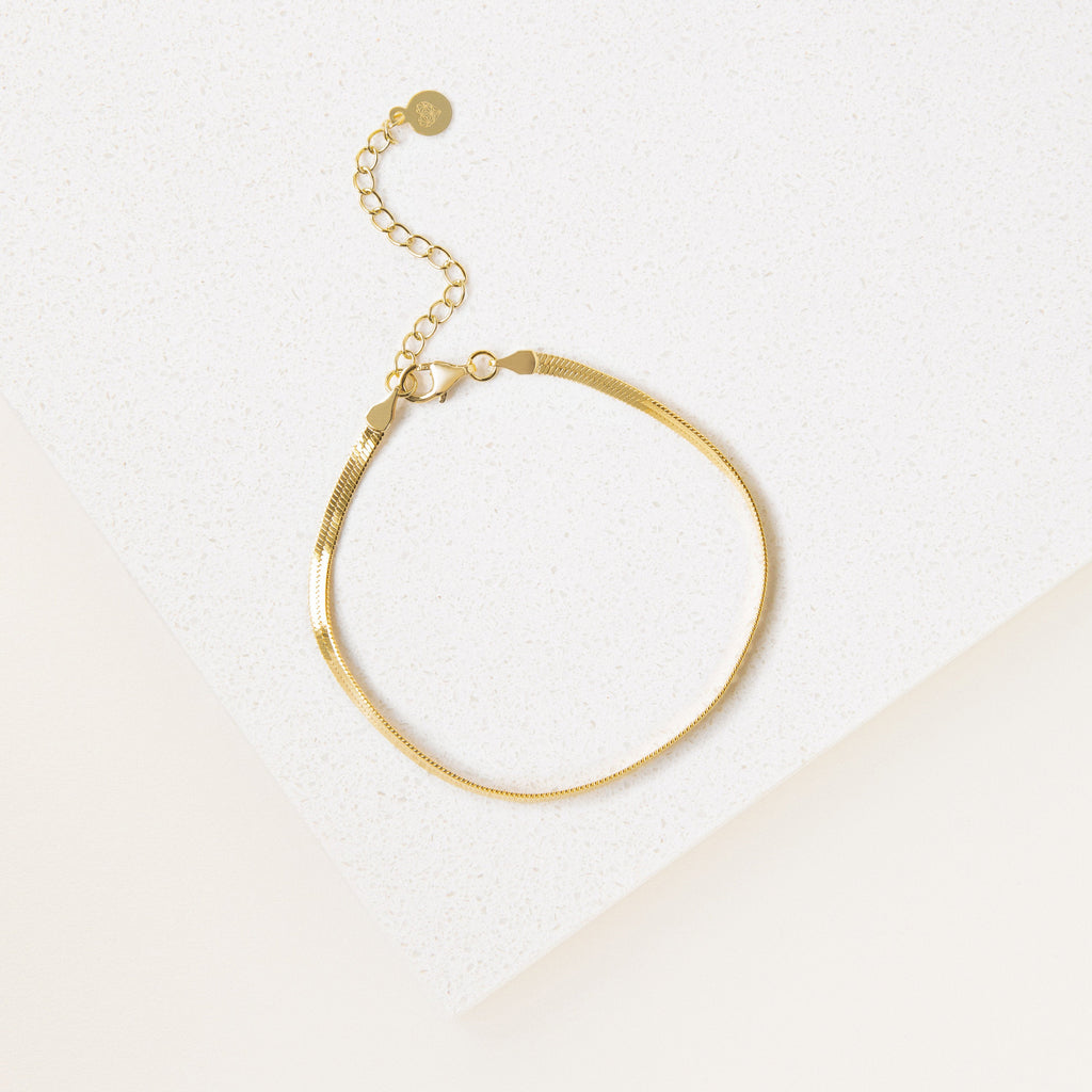 The Essential Bracelet Chains feature a delicate gold chain with polished finishes and an adjustable clasp, shown on a white textured surface.