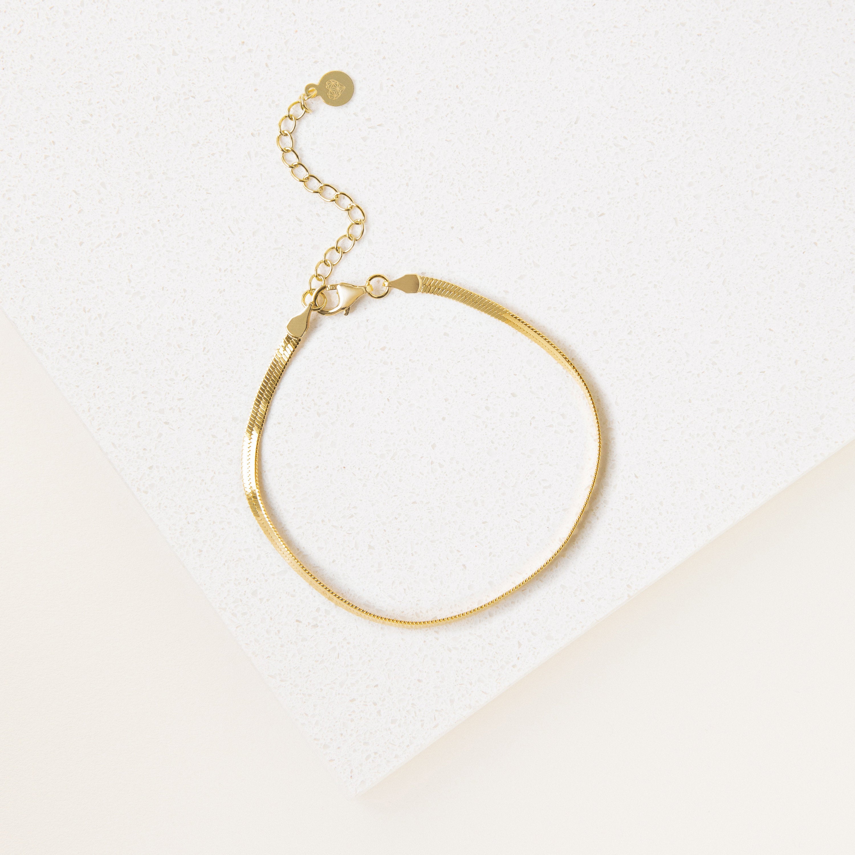 The Essential Bracelet Chains feature a delicate gold chain with polished finishes and an adjustable clasp, shown on a white textured surface.