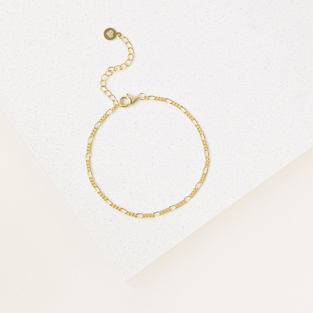 The Essential Bracelet Chains feature a delicate gold chain with an adjustable clasp and polished finish, showcased on a white textured surface.