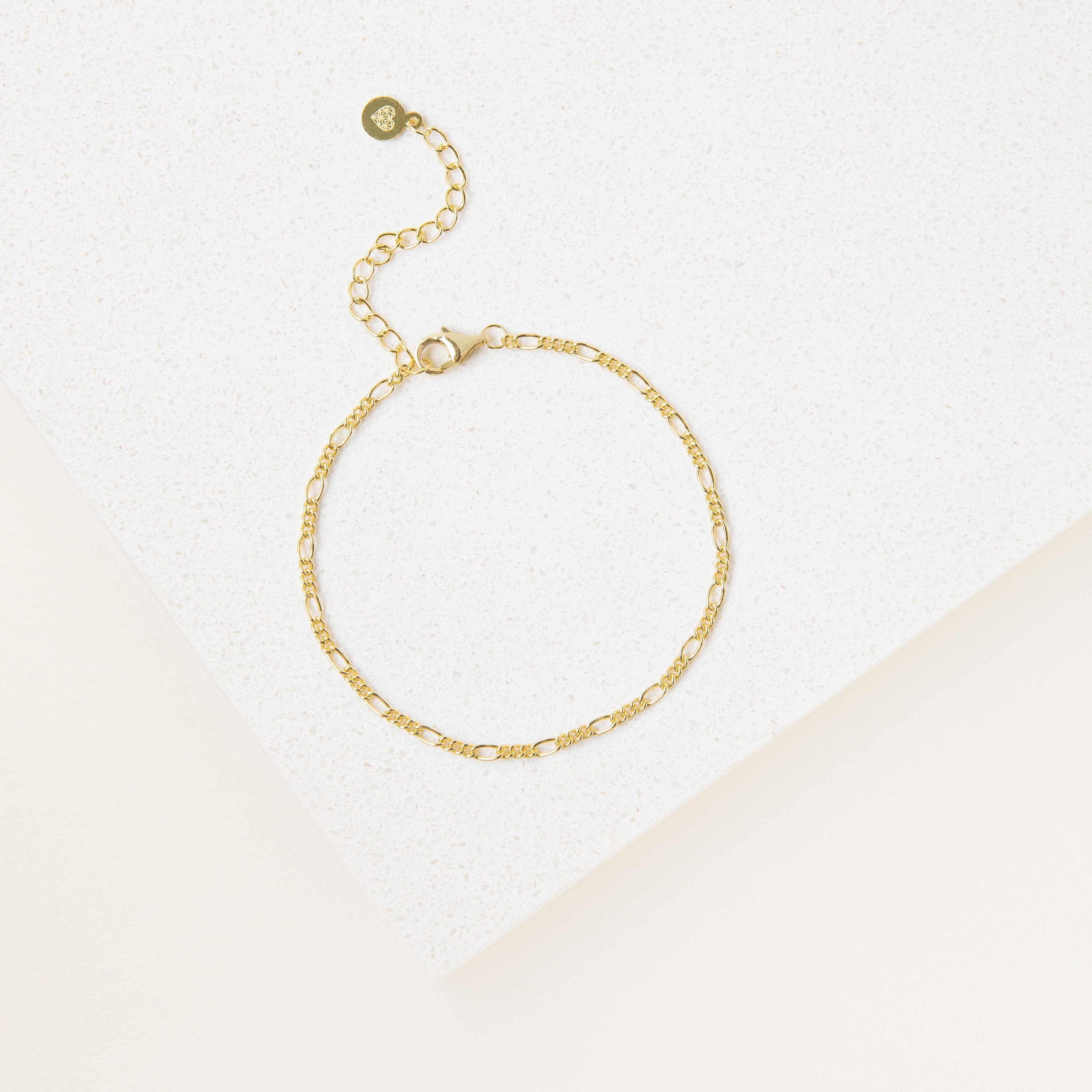 The Essential Bracelet Chains feature a delicate gold chain with an adjustable clasp and polished finish, showcased on a white textured surface.