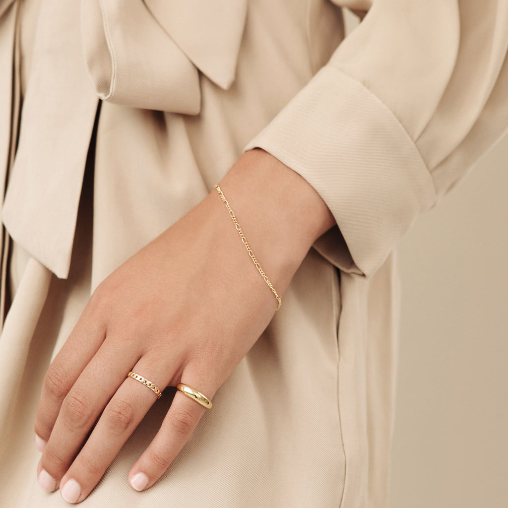 A hand adorned with gold rings and the Figaro Bracelet rests against a beige outfit featuring a bow detail.