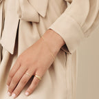 A hand adorned with gold rings and the Figaro Bracelet rests against a beige outfit featuring a bow detail.