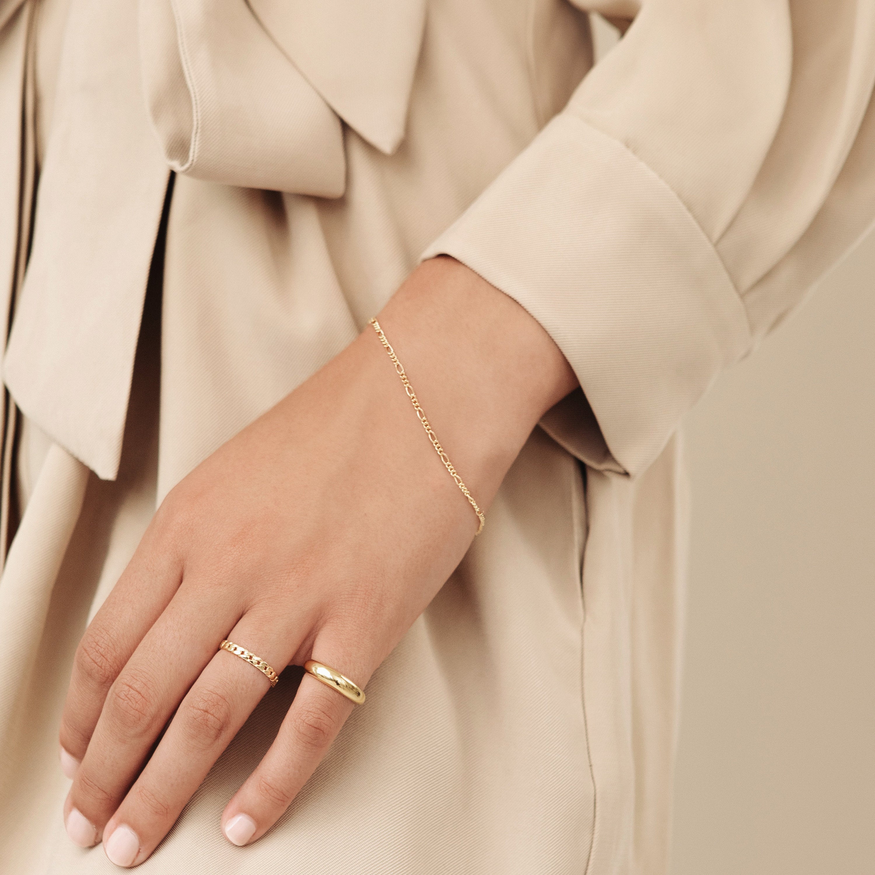 A hand adorned with gold rings and the Figaro Bracelet rests against a beige outfit featuring a bow detail.