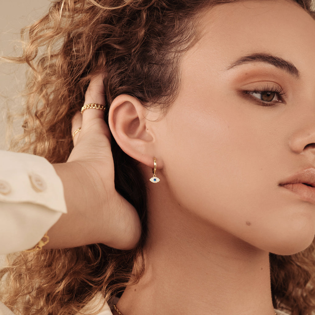 Woman with curly hair gently touching her ear, wearing Iris Huggies—gold earrings featuring an evil eye design for protection.