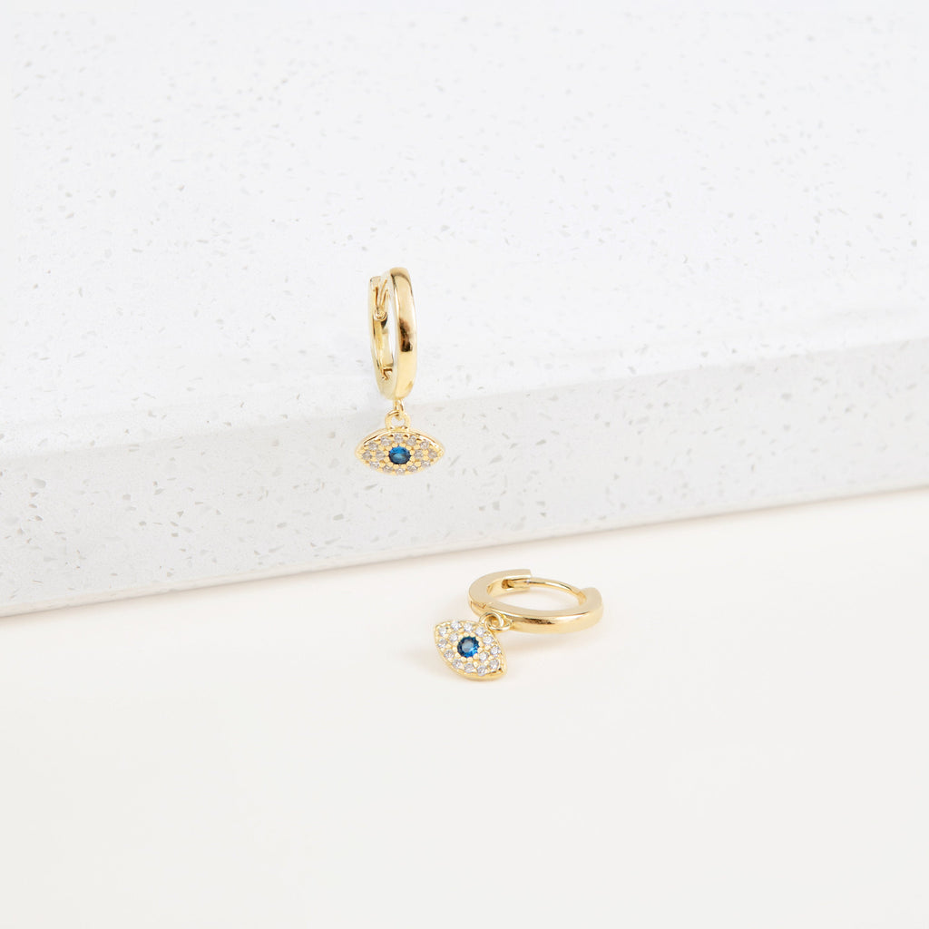 The Iris Huggies are gold earrings featuring blue evil eyes, making them perfect as stylish protection jewelry.