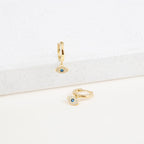 The Iris Huggies are gold earrings featuring blue evil eyes, making them perfect as stylish protection jewelry.