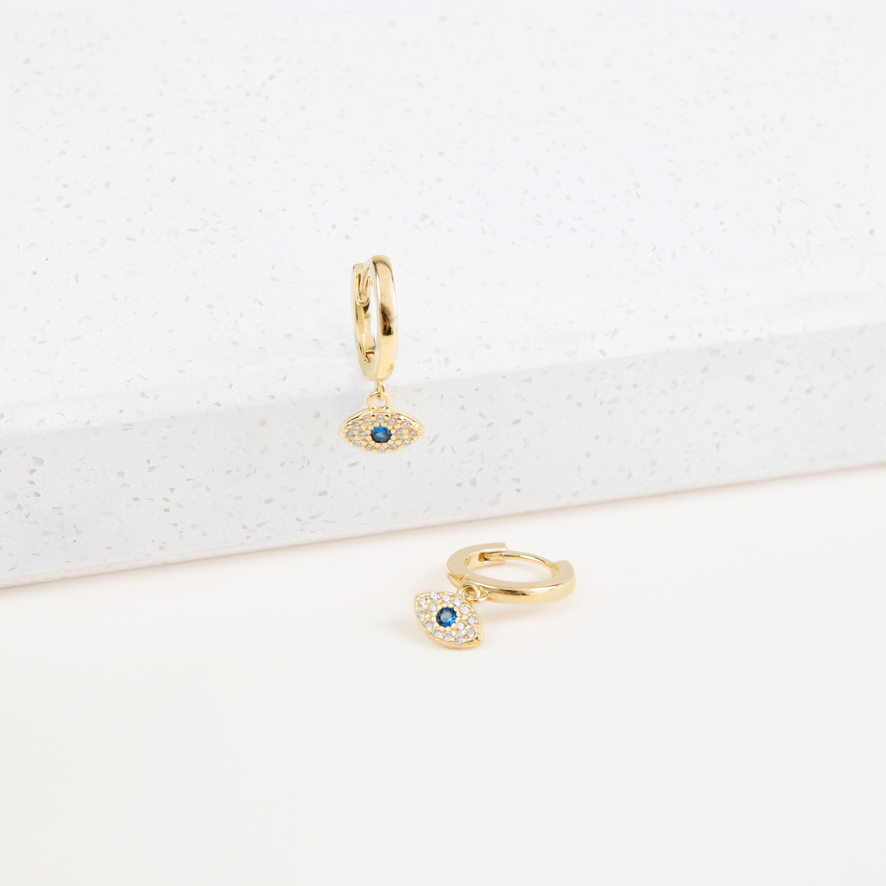 The Iris Huggies are gold earrings featuring blue evil eyes, making them perfect as stylish protection jewelry.