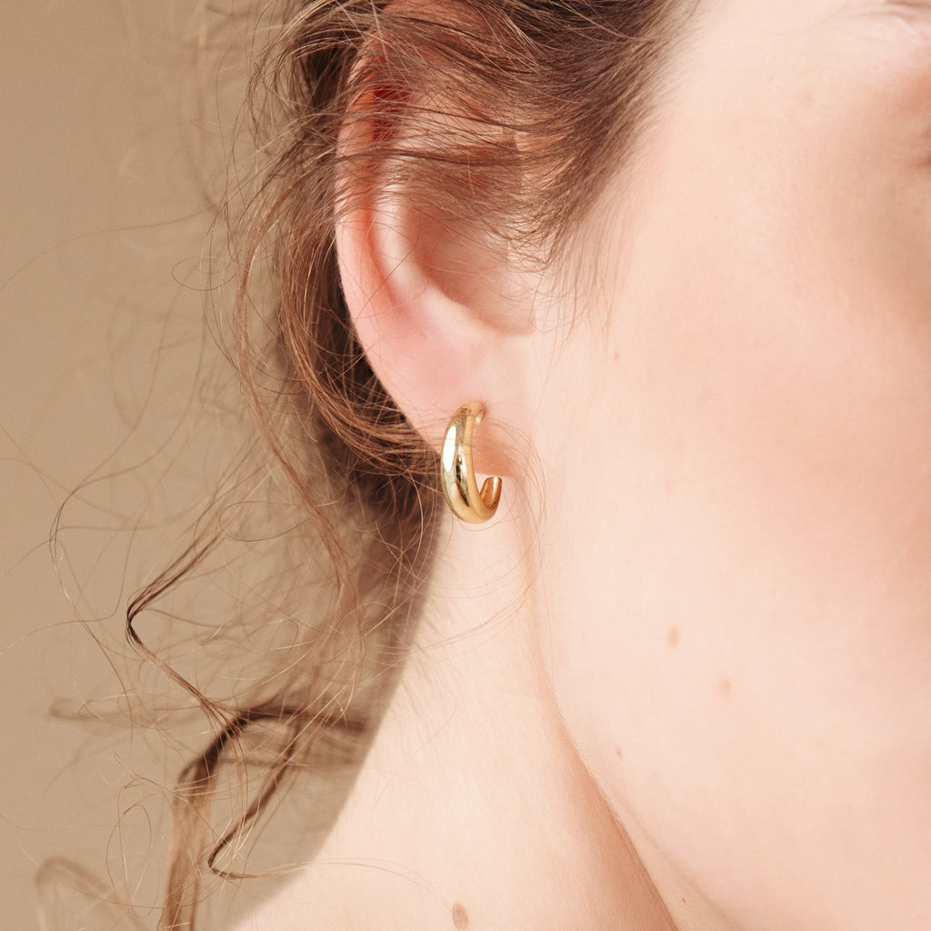 Close-up of a woman's ear wearing small gold Dome Hoops, with her wavy brown hair visible.