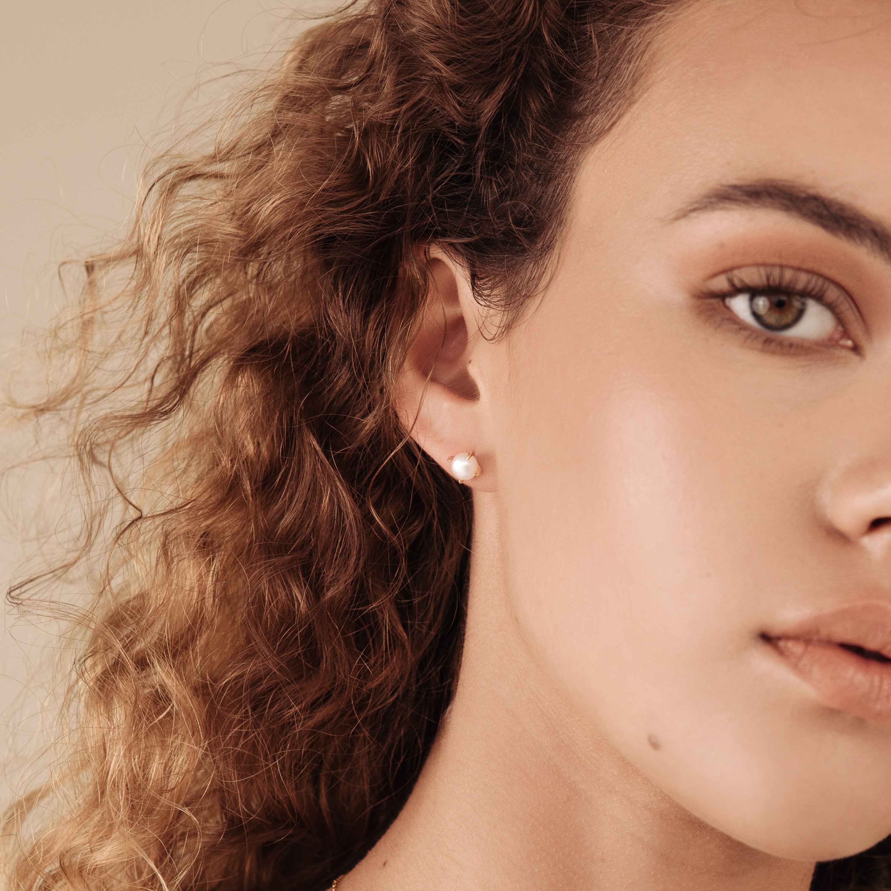 A close-up of a woman with curly hair wearing the Zoe Pearl Studs, highlighting the elegance and modern style of these feminine pearl earrings from the side.